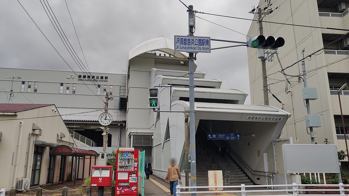 JR 須磨海浜公園站南側,路口號誌桿上掛著藍底白字的「JR須磨海浜公園駅南」路牌,後方灰色站房外牆寫著 JR 須磨海浜公園 SUMA KAIHIN-KOEN STATION,右邊是白色頂棚的步道橋樓梯,左邊有紅色郵筒與自動販賣機