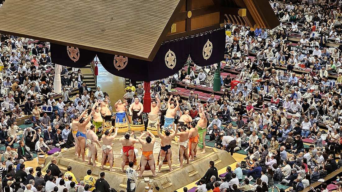 A ring-entering ceremony: more than a dozen wrestlers in colourful embroidered aprons standing in a circle around the edge of the ring with both arms raised, a deep purple valance with gold crests hanging from the roof above, and packed box seats and stands all around