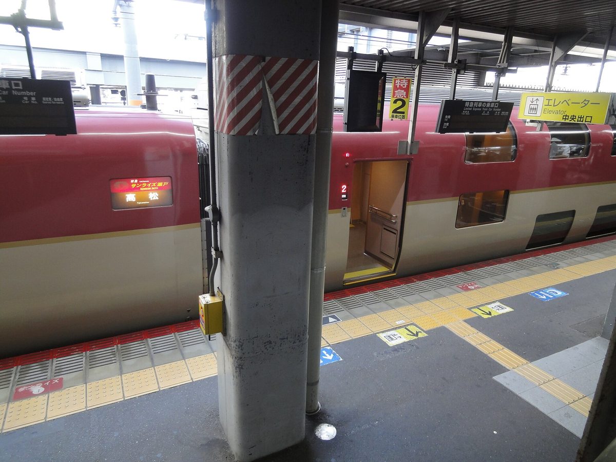 岡山站月台清晨,一列紅白雙色的 Sunrise 停靠:車側的 LED 顯示牌寫著「特急 サンライズ瀬戸 高松」,打開的車門旁亮著紅色數字「2」,月台柱子上掛著黃底「特急 2 号車乗車口」的牌子,右上方是黃色的「エレベーター 中央出口」指示牌