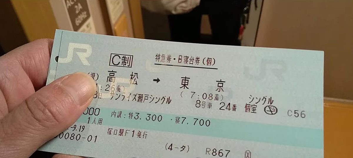 一隻手捏著一張淺藍綠色的 JR 紙本車票,票面印著「特急券・B寝台券(個)」「高松 → 東京(7:08着)」「サンライズ瀬戸シングル」「8号車 24番 シングル 個室」,下方一行「内訳:特3,300・寝7,700」,左側的發車時刻被拇指擋住一半