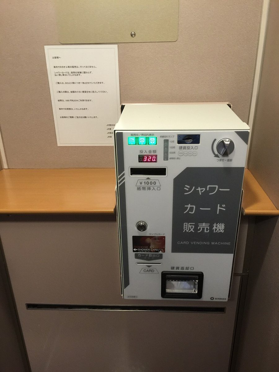 車廂牆邊一台白色的「シャワーカード販売機」,面板上有投幣口、只收一千圓的紙鈔口、顯示 320 的投入金額紅色數字,下方是取卡口與退幣口;左上方牆上貼著一張日文公告