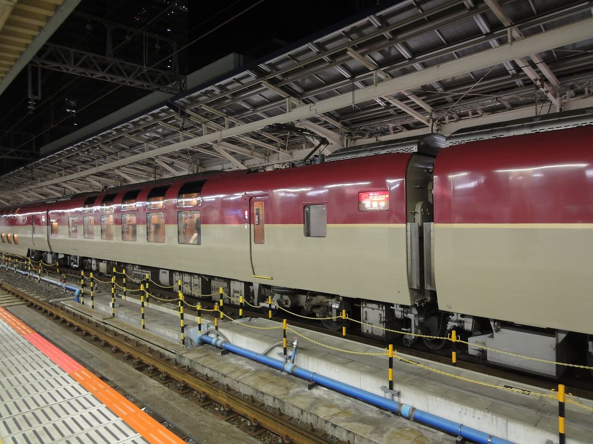 夜裡的東京站月台,鋼骨雨棚下停著一列 Sunrise:車廂上半紅色、下半米白,車窗分上下兩排、裡面亮著暖黃色的燈,車門上方的小螢幕亮著紅字,月台邊緣是黃黑相間的護欄與藍色水管