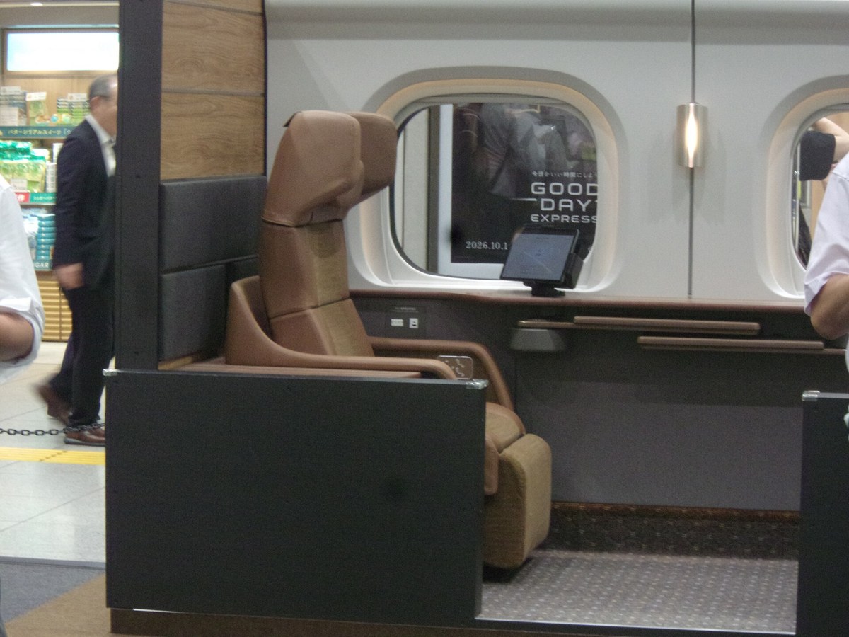 A full-size Supreme Class Cabin mock-up standing on a station floor: a section of white car body with two rounded windows, and inside a tall brown leather seat with a headrest plus a lower seat behind it, a wooden counter with two rails under the window, a small tablet on a stand, and a black poster visible through the glass reading GOOD DAY EXPRESS 2026.10.1; a station shop and a passer-by appear at the left edge