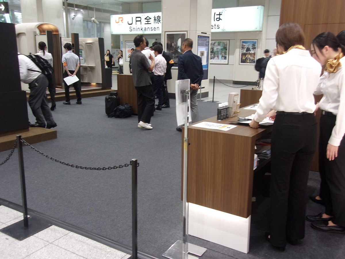 The Supreme Class mock-up event in a station concourse: on the left the white car-body mock-up with several people looking inside and a uniformed staff member in an orange armband holding papers; behind it an illuminated ticket-office sign reading JR全線 and Shinkansen; on the right a wooden reception counter with leaflets and bottled water staffed by two women in scarves; a chain-and-post barrier runs across the foreground