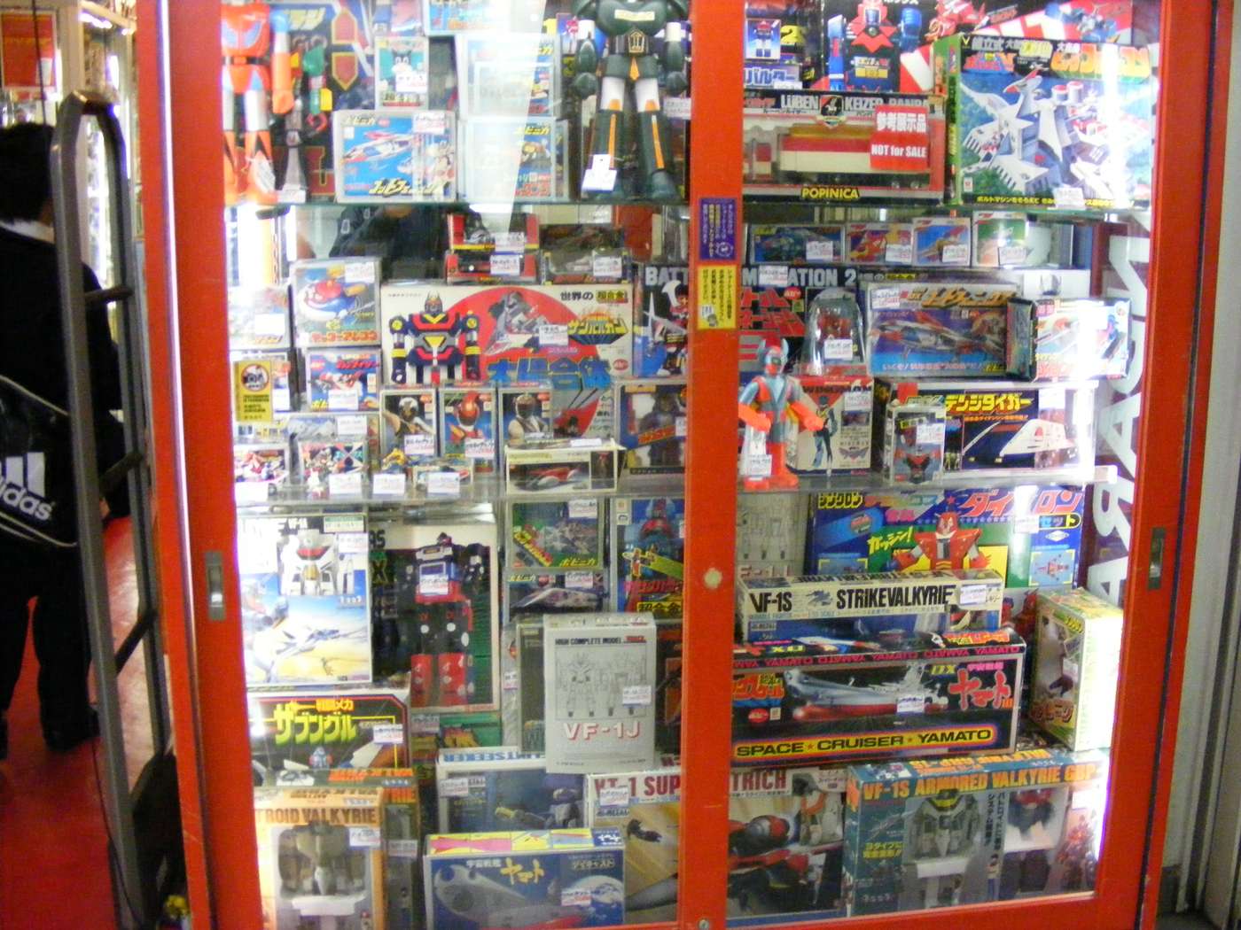 A glass display case in a secondhand toy shop packed with boxed robot model kits and out-of-print figures, each individually tagged