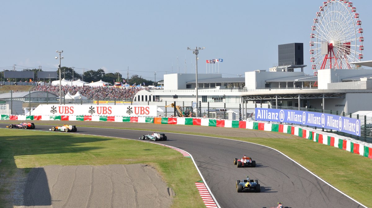 2012 年 F1 日本大獎賽中，六台賽車排成一列通過鈴鹿的連續彎，後方是白色的賽道建築與坐滿觀眾的看台，右上角是紅白色的大摩天輪
