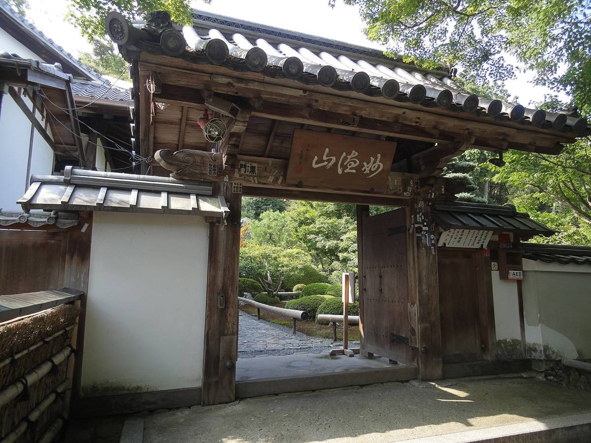 鈴蟲寺(華嚴寺)的山門:瓦頂木造門上掛著「妙徳山」匾額,門板貼滿千社札,右側掛著寫有見學時間與志納料的木牌,門內看得到石板路與修剪過的庭木