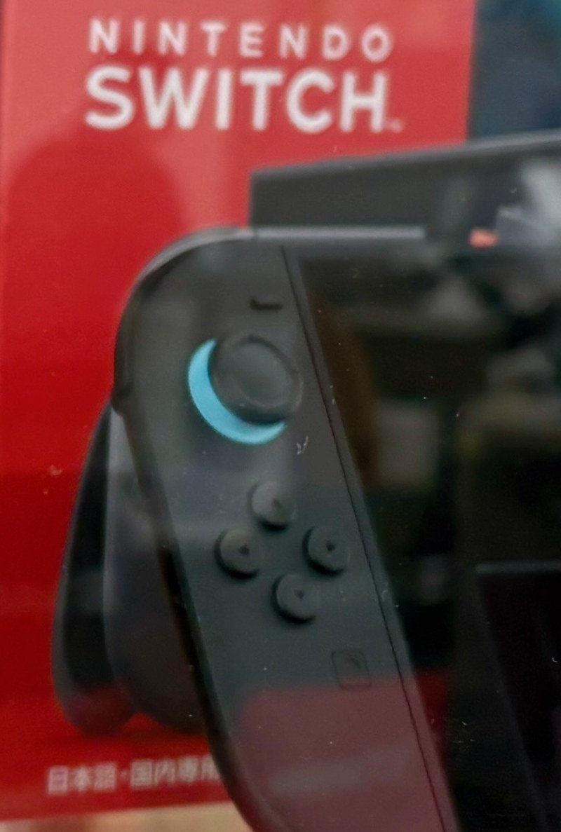 隔著玻璃拍的 Nintendo Switch 2 紅色包裝盒正面，盒子左下角有一行白色小字「日本語・国内専用」，最後一個字被擺在盒子前面的主機擋住一部分