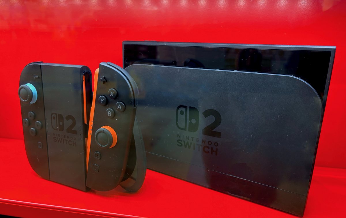 紅色展示櫃裡的 Nintendo Switch 2：主機插在印著 Switch 2 標誌的黑色底座上，左邊是裝在握把上的一對 Joy-Con 2，搖桿底下一藍一橘