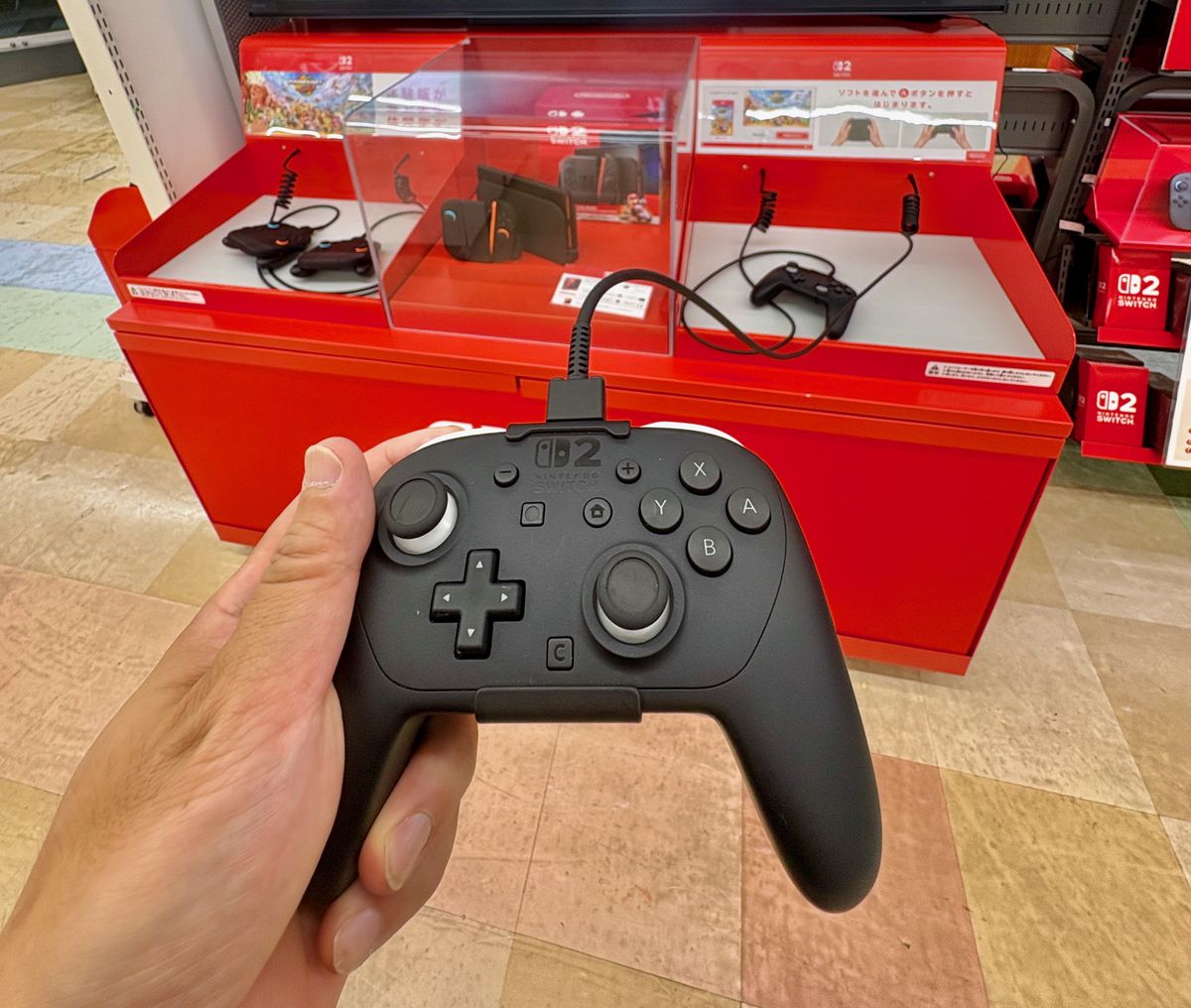 一隻手握著黑色的 Switch 2 Pro 控制器，上緣接著一條防盜線，右搖桿左下方有一顆方形的 C 鍵；後方是紅色的 Switch 2 試玩台，台上放著 Joy-Con 2 與另一支 Pro 控制器