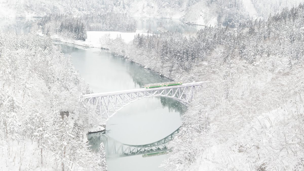 2018 年 1 月大雪中的第一只見川橋梁:從高處俯瞰,拱橋與兩岸樹林幾乎全白,橋上一列綠白色的兩節列車,拱橋的倒影映在灰綠色的河面上