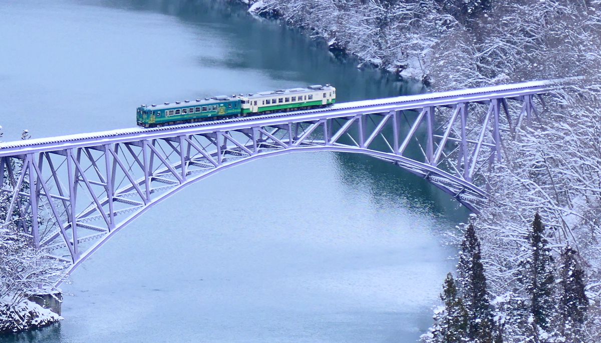 列車通過第一只見川橋梁的近景:橋面積雪的淡紫色拱橋上,一節深綠色與一節白底綠帶的柴油車連在一起行駛,橋下是青綠色的只見川