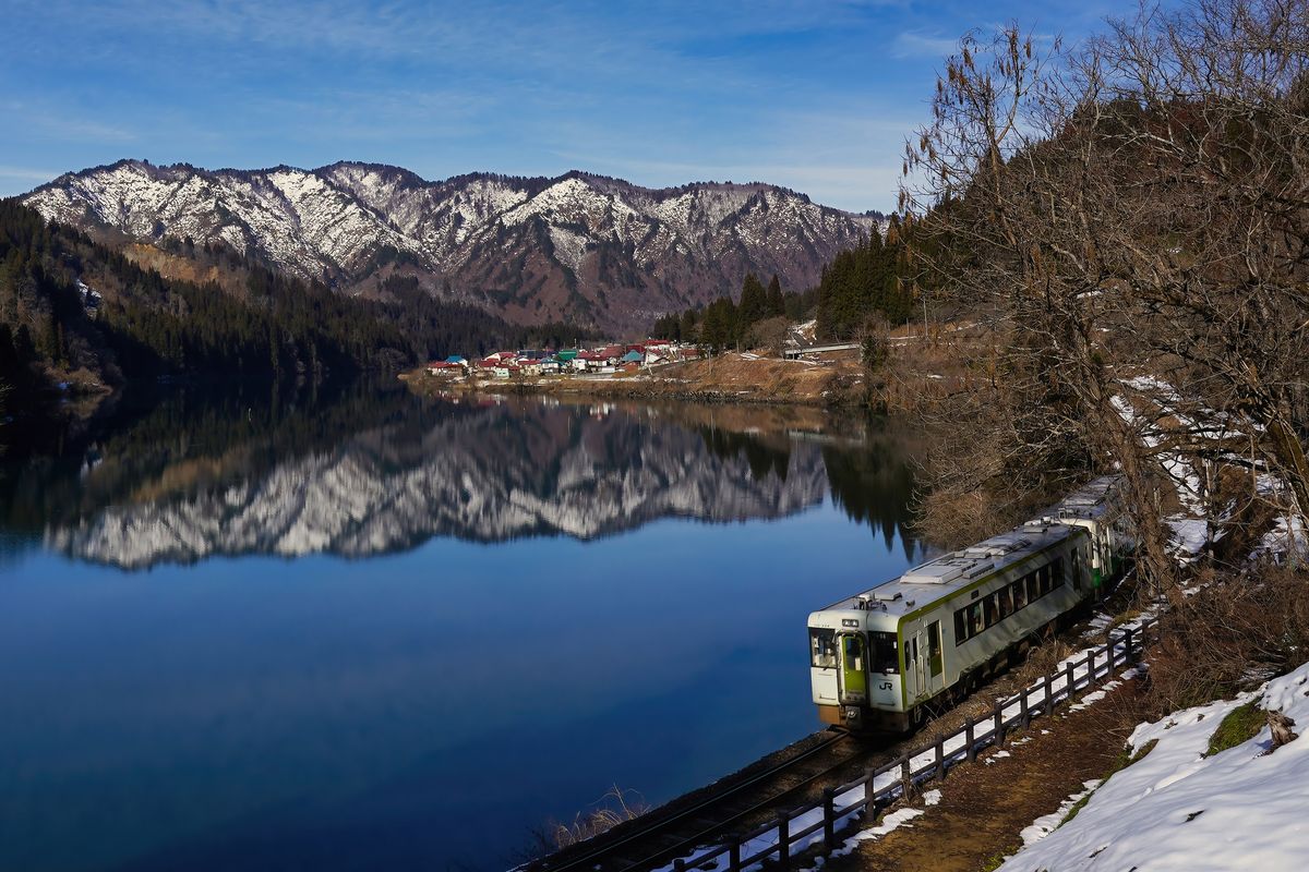 2024 年 1 月只見線的キハ110 型列車:白底配黃綠色車門的兩節柴油車沿著只見川邊的軌道行駛,平靜的河面映出對岸積雪的山,遠處河邊有一小片紅藍屋頂的聚落