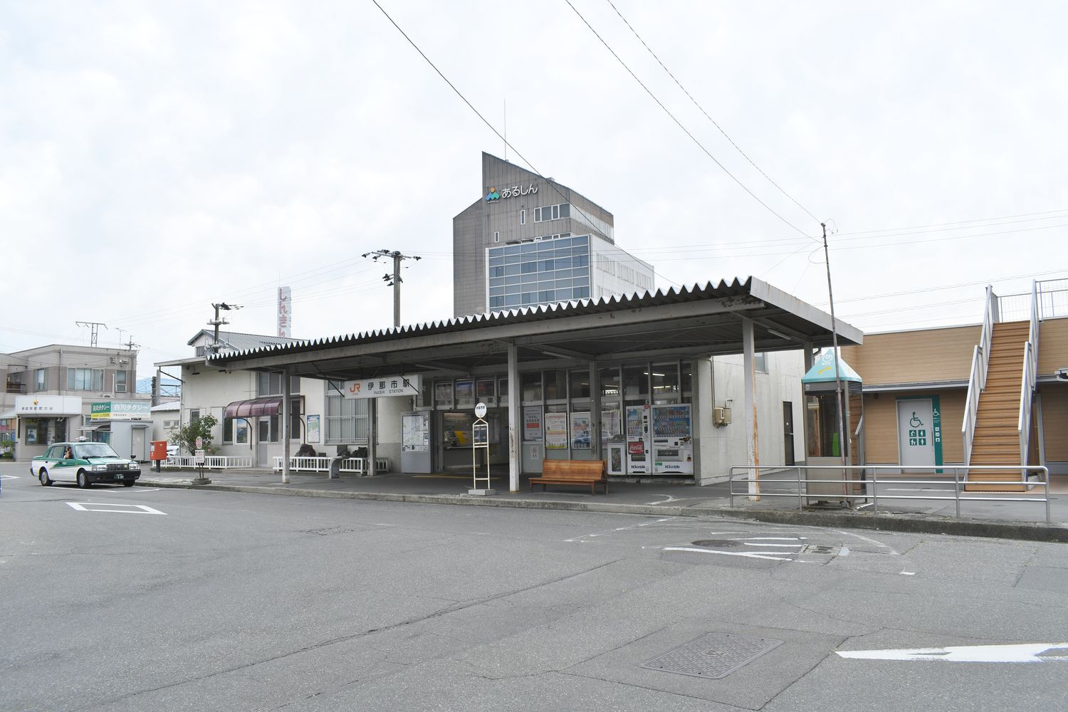JR 飯田線伊那市駅的站舍:一棟平房式的小車站,屋簷下掛著 JR 的藍色站名牌與「伊那市駅」字樣,站前停著一台計程車,右側有幾台自動販賣機