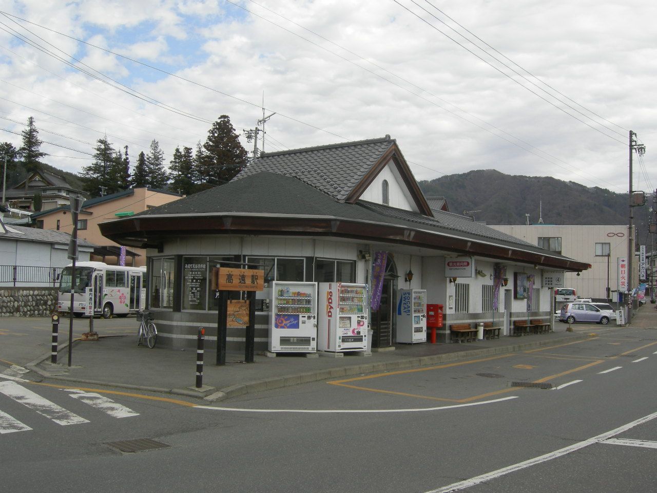 JR バス高遠駅:一棟灰瓦屋頂的平房,門口立著寫著「高遠駅」的木牌與觀光案內所的招牌,牆邊排著三台自動販賣機,左側停著一台小型巴士