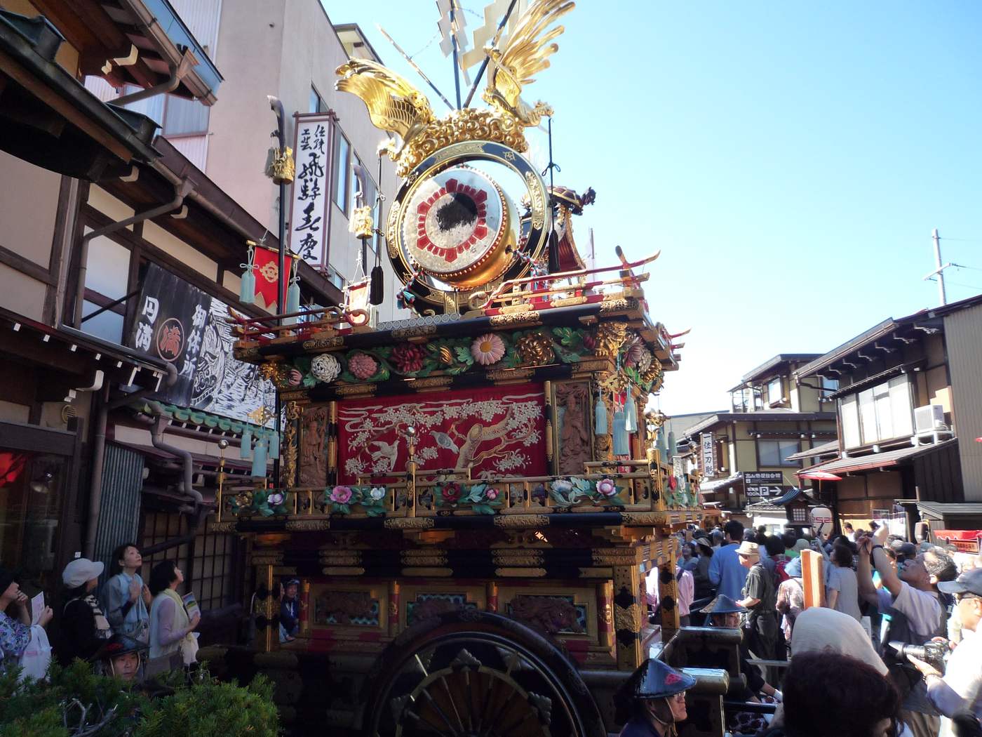 An autumn Hachiman Festival yatai float parked in a Takayama street, gilt phoenix and carved panels catching the sun, onlookers on both sides