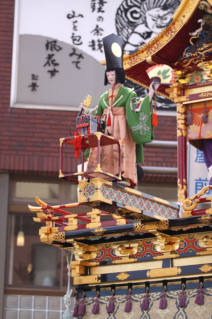The Sanbaso karakuri puppet in a green robe holding a fan, standing on the extended bridge mechanism of its float