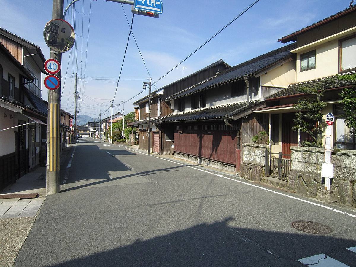 A street in the Takeda castle town: traditional dark-timber lattice-fronted townhouses and low stone walls along one side, a bus stop pole and a 40 km/h speed sign at the kerb, with mountain ridges closing the far end of the street