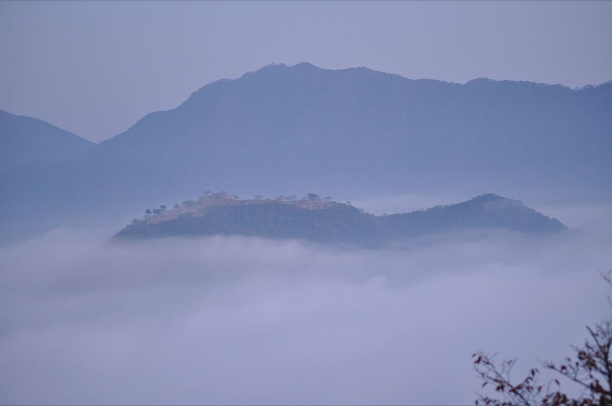 Dawn view from Ritsuunkyo: the ridge holding Takeda Castle ruins riding above a white sea of clouds, with faint lines of stonework along the summit and layered mountain silhouettes behind