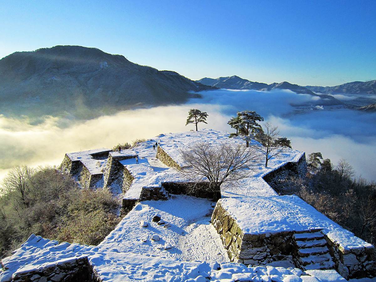 Snow-covered enclosures and stone walls of Takeda Castle ruins with a few pines, several visitors walking in the distance, and a sea of clouds filling the valley beyond under a deep blue sky