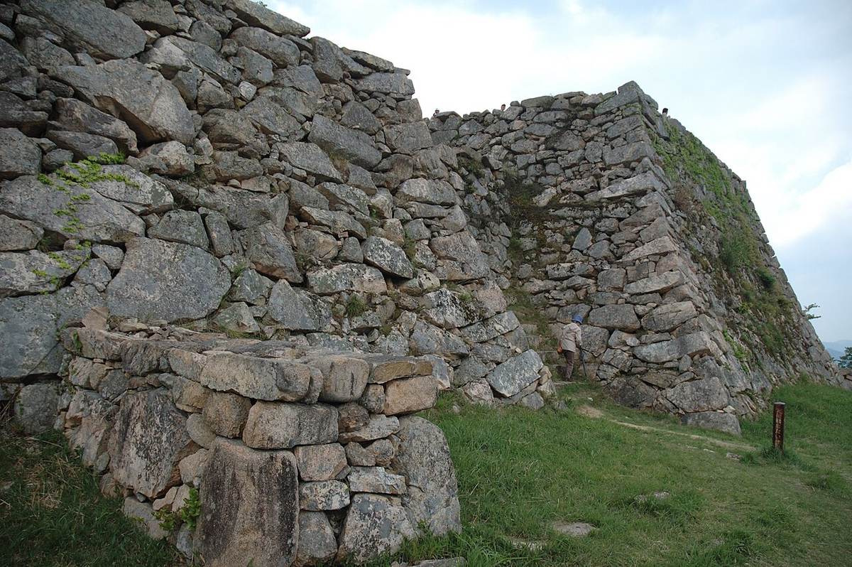Close view of nozura-zumi stonework in the honmaru area of Takeda Castle: rough, barely worked stones of wildly different sizes stacked into high walls and a sharp corner, grass at the base, a single visitor standing beneath for scale