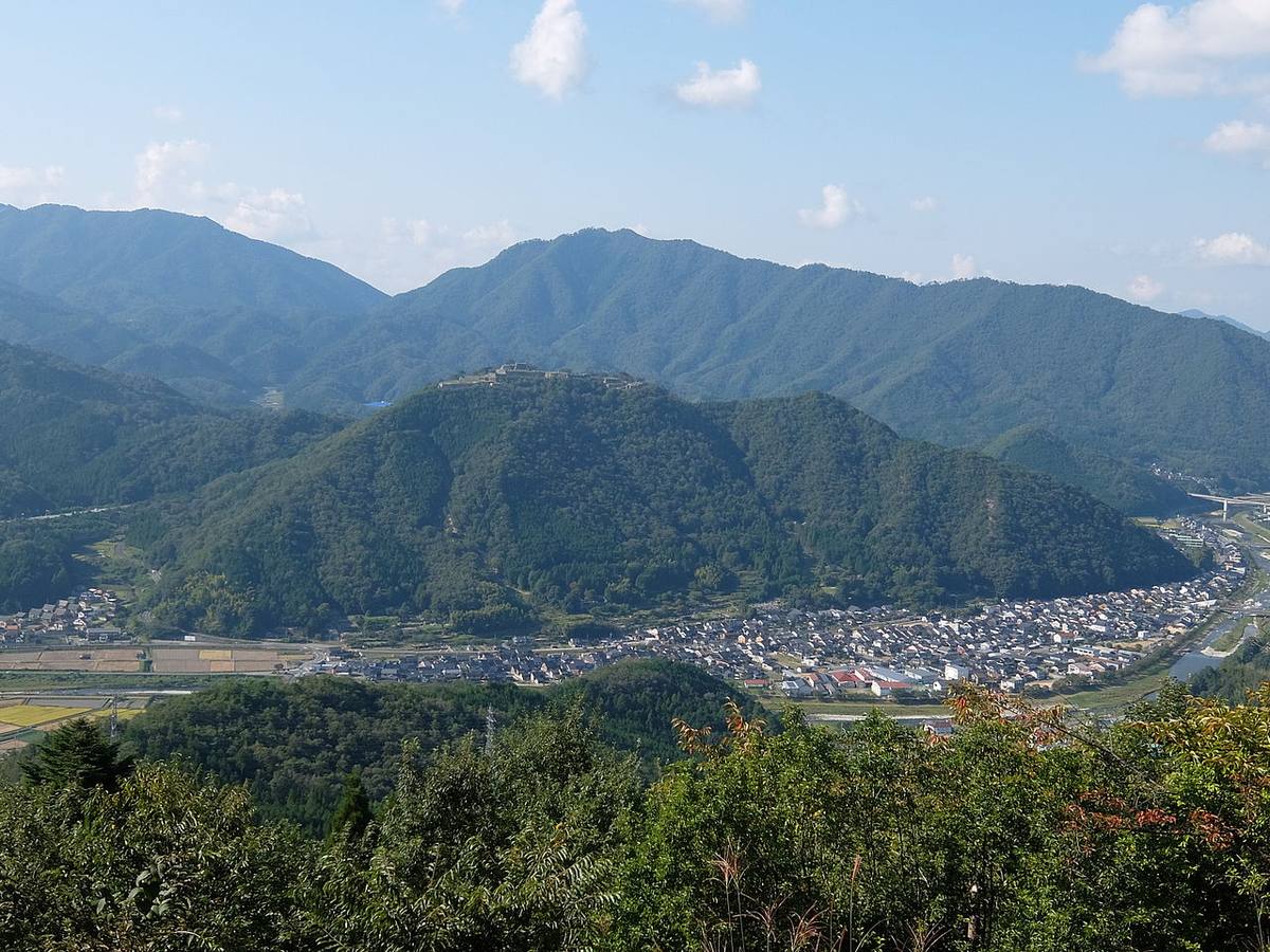 View across the valley from the Ritsuunkyo side: the stone ramparts of Takeda Castle clearly visible along the summit opposite, the town of Takeda and its rice fields on the valley floor, and the river and highway to the right