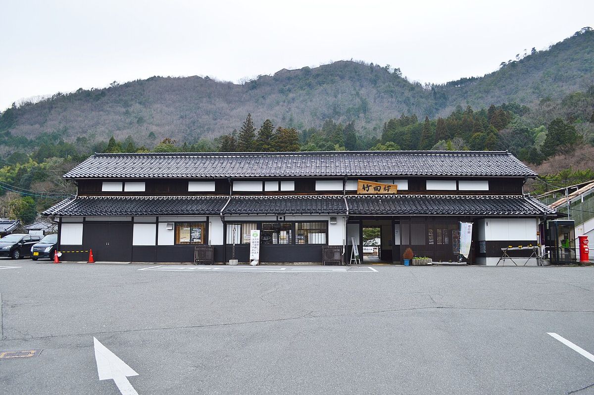 Exterior of JR Takeda Station: a black-and-white traditional-style building with a wooden signboard reading Takeda Station under the eaves, an empty forecourt in front and the castle mountain rising behind