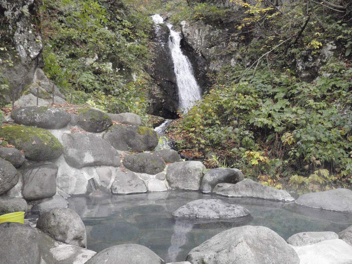 A rock-lined open-air hot spring pool in the mountains, steam rising off the surface and a boulder breaking the water in the middle, a narrow waterfall dropping down the cliff behind, green shrubs and scattered dead leaves around, and a yellow plastic bucket at the lower left