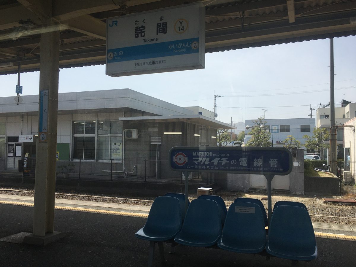 JR 詫間站的月台：屋簷下掛著白底藍條的站名牌，寫著詫間、Takuma、站號 Y14，左右分別是往みの與往かいがんじ，對面是一層樓的灰白色站房，月台上有一排藍色塑膠椅