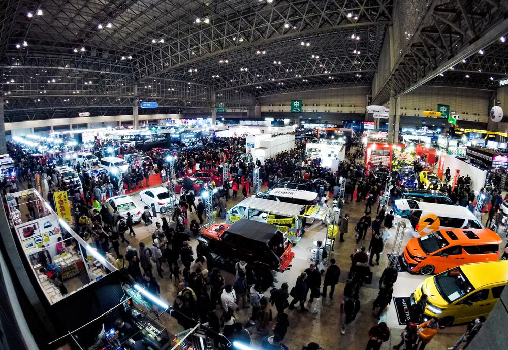 用廣角從展館高處往下拍的車展週六中午:格狀鋼骨天花板下,整個展館的走道擠滿人,攤位之間幾乎看不到空地,右前方是橘色和黃色的改裝廂型車