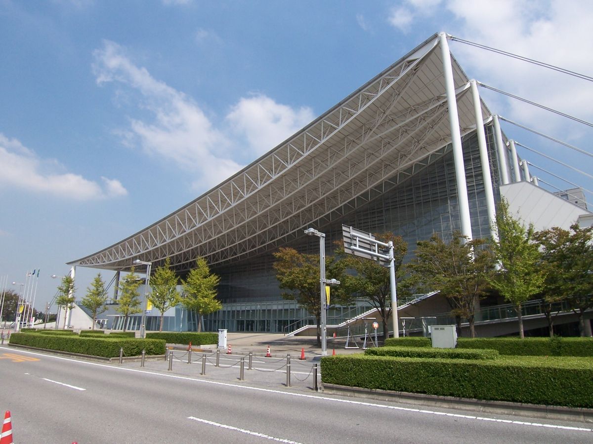 幕張メッセ國際展示場的外觀:一整片向上斜挑的鋼骨大屋頂,屋簷下是玻璃立面的展館,前面是車道與修剪整齊的綠籬,晴天