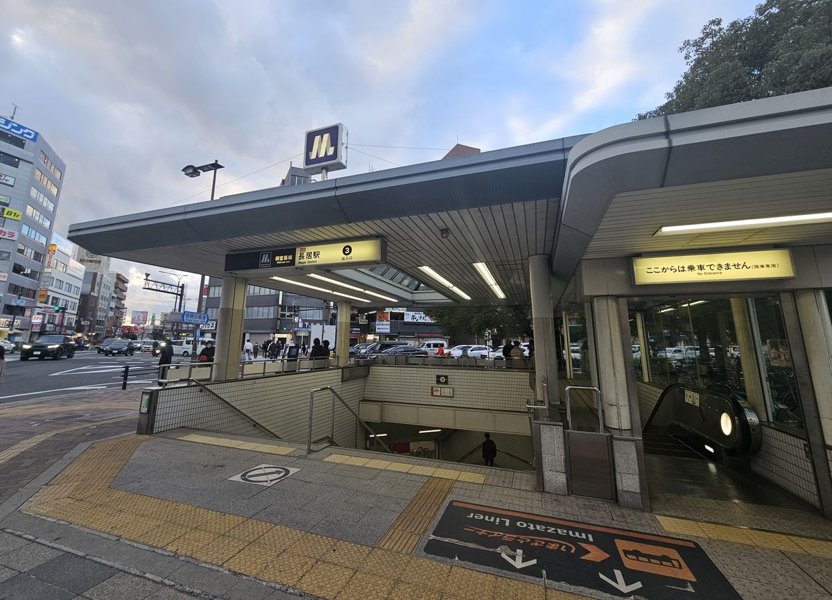 Osaka Metro 長居站 3 號出入口外觀:灰色頂棚下掛著黃底燈箱「御堂筋線 長居駅 ③ 出入口」,頂上有 Osaka Metro 的 M 字標誌,左邊是往下的樓梯、右邊是手扶梯,左後方路口立著藍色路名牌「長居公園通 Nagai Koen-dori」,遠處行人很小