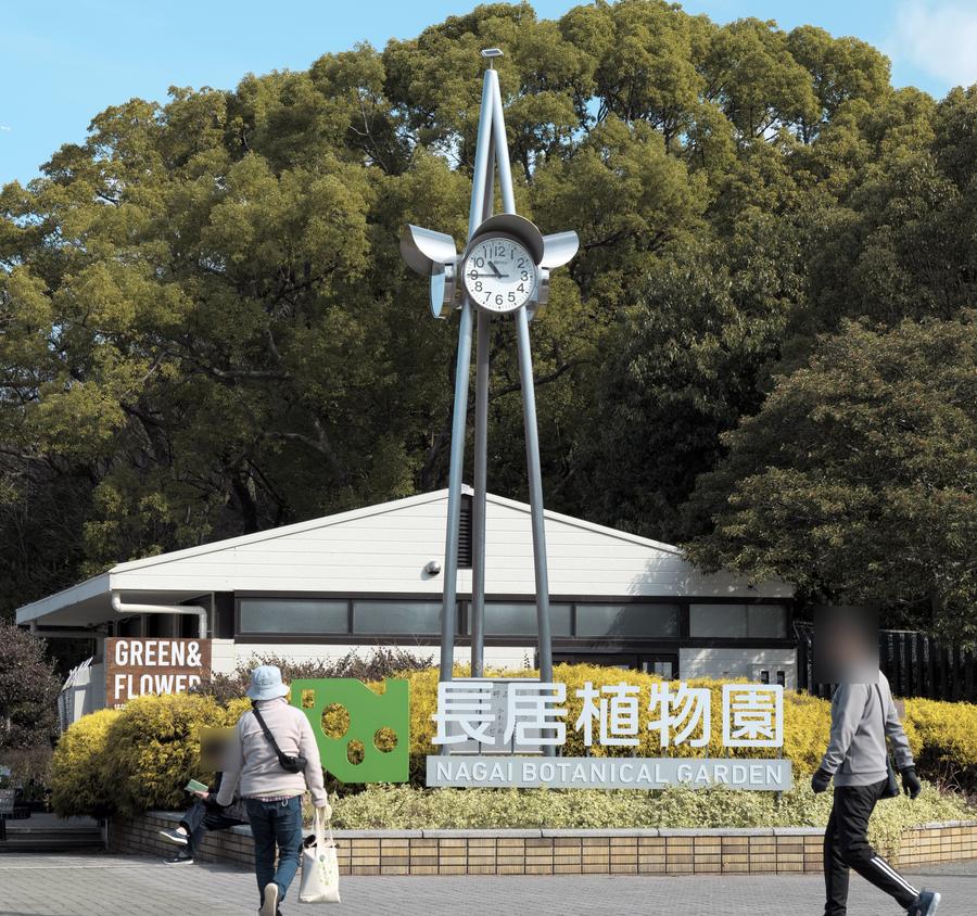 長居植物園的園名標示:花壇上白色立體字「長居植物園 NAGAI BOTANICAL GARDEN」與綠色植物園標誌,後方一座銀色時鐘塔、一棟白色平房,再後面是一大片濃密的樹林;前景有幾位路過的遊客,臉部已模糊