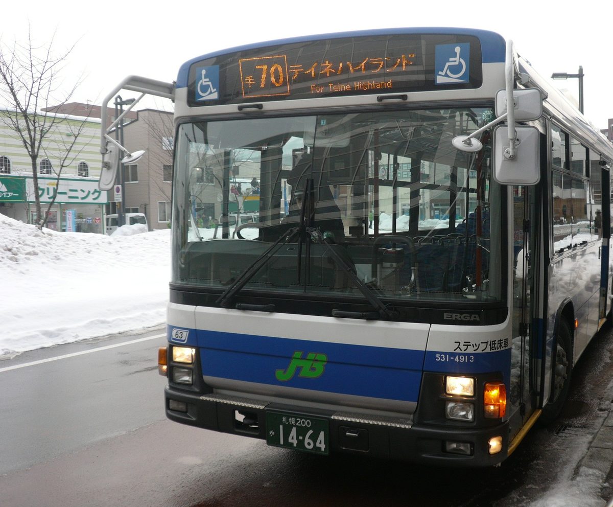 A white-and-blue JR Hokkaido Bus standing at a snowed-in bus shelter, its front LED sign reading in orange 'te-70 テイネハイランド / For Teine Highland', with the green JHB logo and the words ステップ低床車 (low-floor bus) along the skirt and ploughed snow banked up beside the road