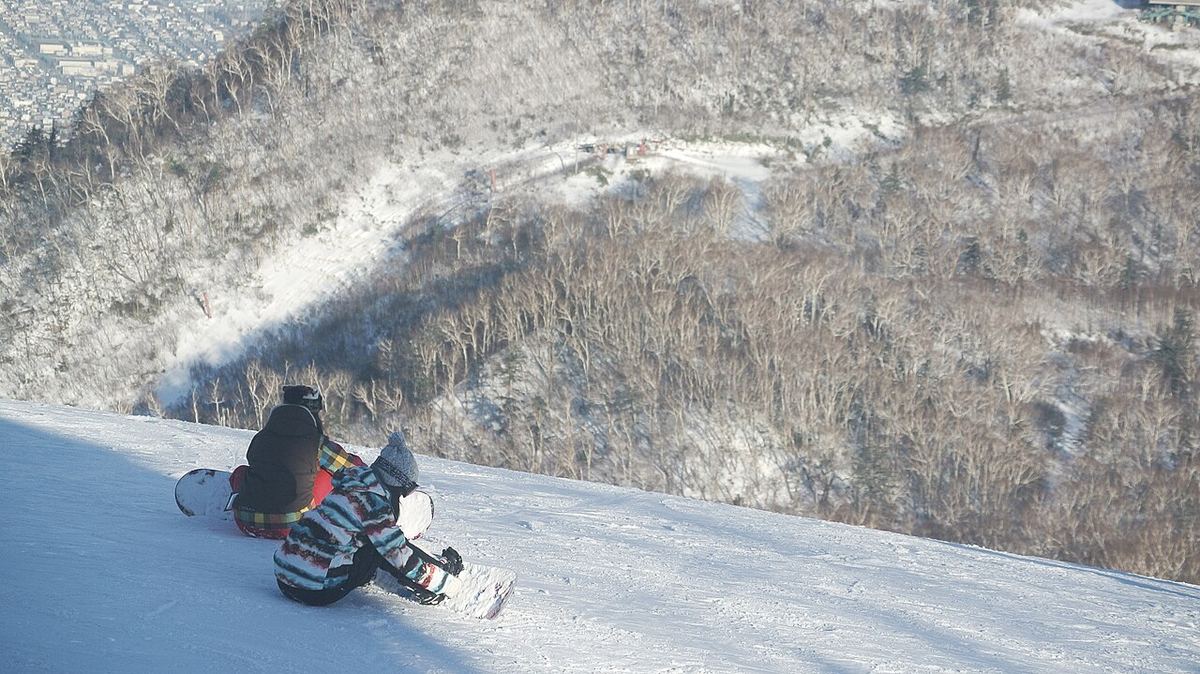 Two snowboarders sitting with their backs to the camera on the edge of a groomed run at Sapporo Teine, a wooded valley below and dense Sapporo housing visible at the top left