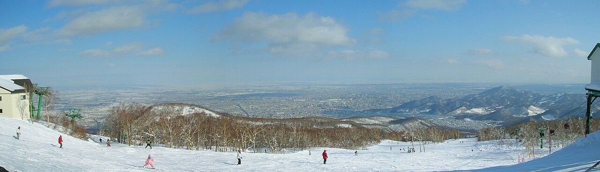 Ultra-wide panorama from the upper runs at Mt. Teine, skiers scattered across the groomed slope with a chairlift on each side and the Sapporo cityscape and plain stretching to the horizon
