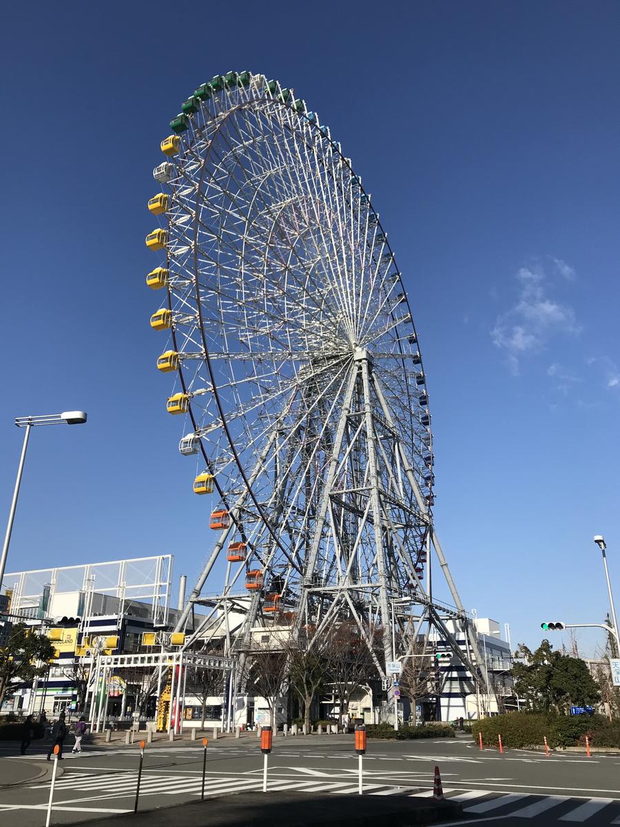晴天下仰拍天保山大摩天輪,白色鋼架上掛著黃、綠、橘色車廂,左下是天保山ハーバービレッジ的建築與 LEGOLAND 招牌,前景是路口斑馬線