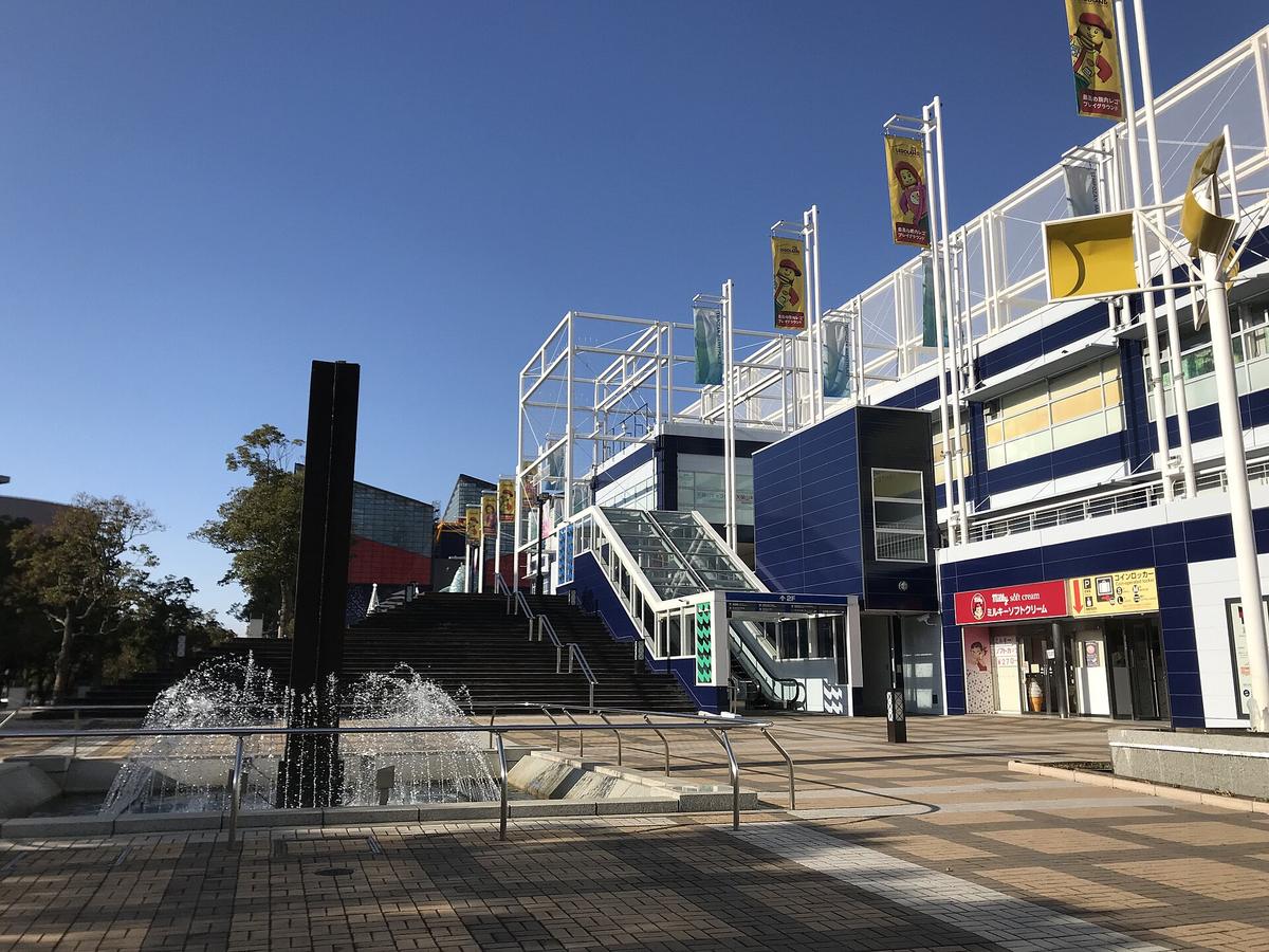 天保山マーケットプレース的藍白色建築與通往二樓的大階梯、手扶梯,前景是噴水池,階梯左上方露出海遊館的紅色外牆與玻璃帷幕,晴天無人