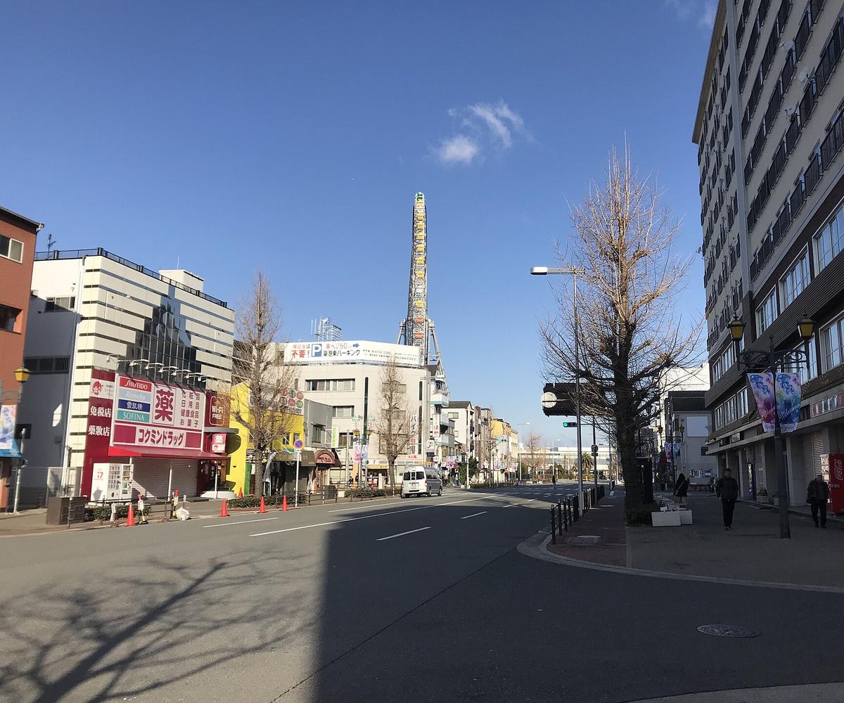 大阪港站附近一條寬闊的街道,兩側是藥妝店與高樓,路的盡頭從建築後方露出天保山大摩天輪的側面,冬天的行道樹沒有葉子,晴天