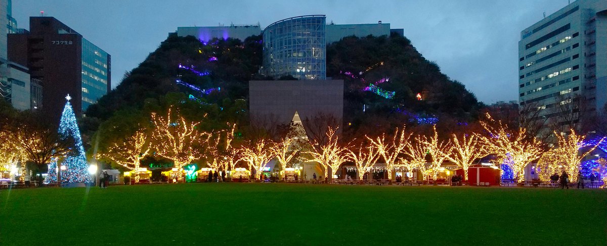 2023 年 12 月傍晚的天神中央公園：大草坪盡頭一排纏著暖黃色燈串的樹，樹下是一間間亮燈的攤位與長桌，左邊一棵藍白色燈飾的聖誕樹，後方是外牆階梯狀種滿樹、綴著紫色與藍色燈的 ACROS 福岡大樓