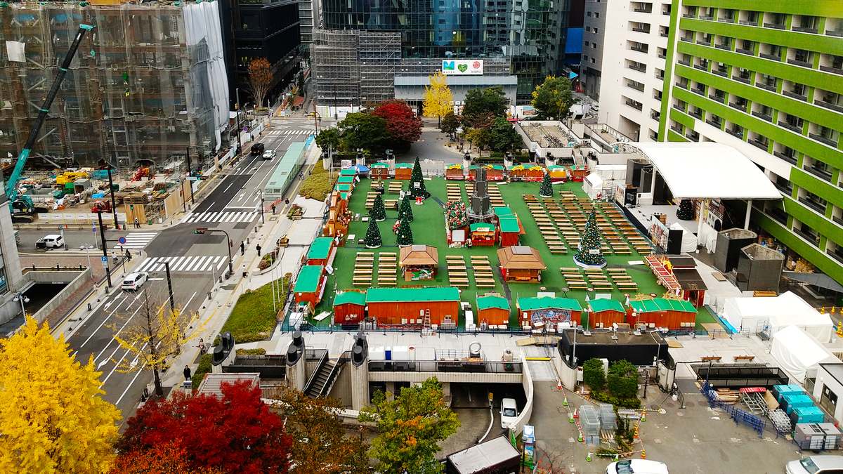 2025 年 11 月平日下午從高樓俯看天神的福岡市役所西側ふれあい廣場：綠色地墊上排著一圈綠屋頂的紅褐色木屋攤位，中間是幾棵聖誕樹與成排的空長桌，右側有白色弧形頂棚的舞台，旁邊是綠色橫條外牆的大樓，左側是馬路與施工中的大樓，前景有黃色銀杏與紅葉