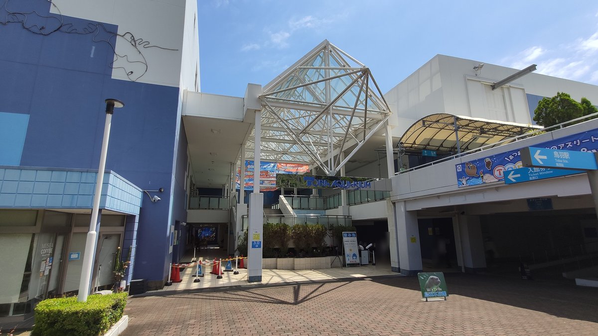鳥羽水族館入口廣場:玻璃鋼架屋頂下是 TOBA AQUARIUM 招牌和通往館內的通道,右上方兩塊藍色指標寫著「鳥羽駅」與「入館券 Ticket Window 券売口」,箭頭都往左指