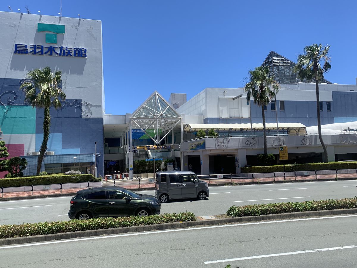 馬路邊的鳥羽水族館正面:左邊藍白相間的大樓頂掛著綠色 T 字標誌和「鳥羽水族館」,中間是玻璃鋼架的入口屋頂,前面種著棕櫚樹