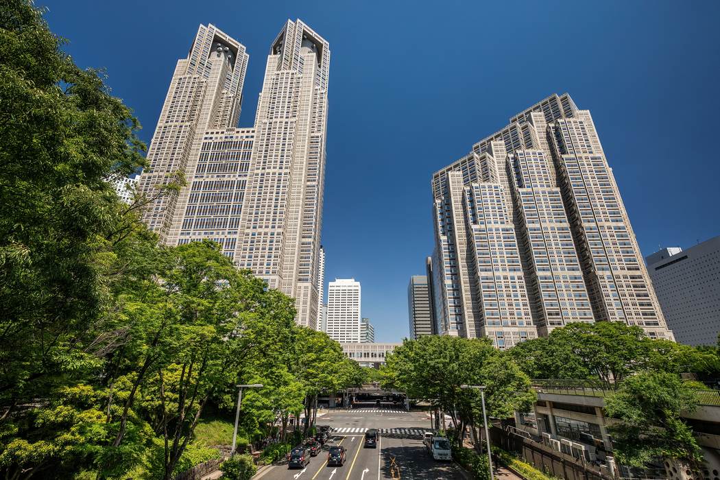 晴天下一條兩旁種滿綠樹的道路直直通往兩棟高樓，左邊是頂部分成雙塔的第一本廳舍，右邊是層層退縮的第二本廳舍，路上有幾輛車