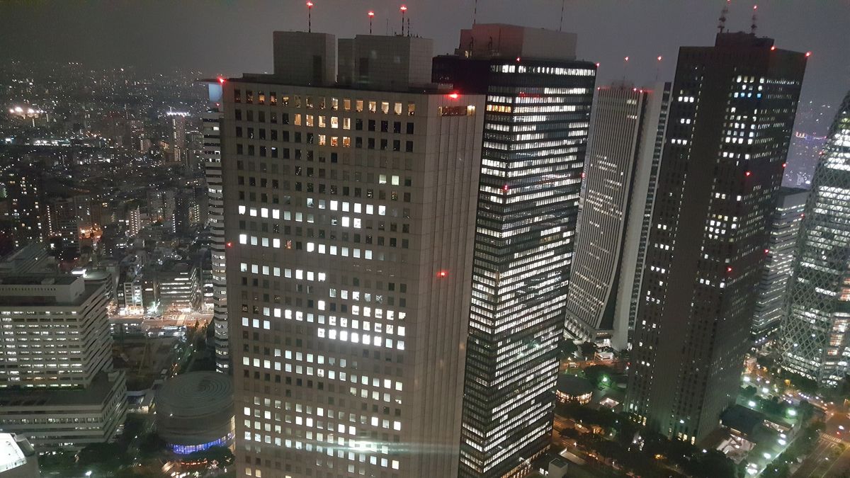 夜間從高處斜看一排西新宿摩天樓，近處一棟方正的淺色大樓窗戶半亮，旁邊幾棟深色玻璃高樓亮著橫條窗光，樓頂閃著紅色航空燈，左側遠方是一片城市燈海