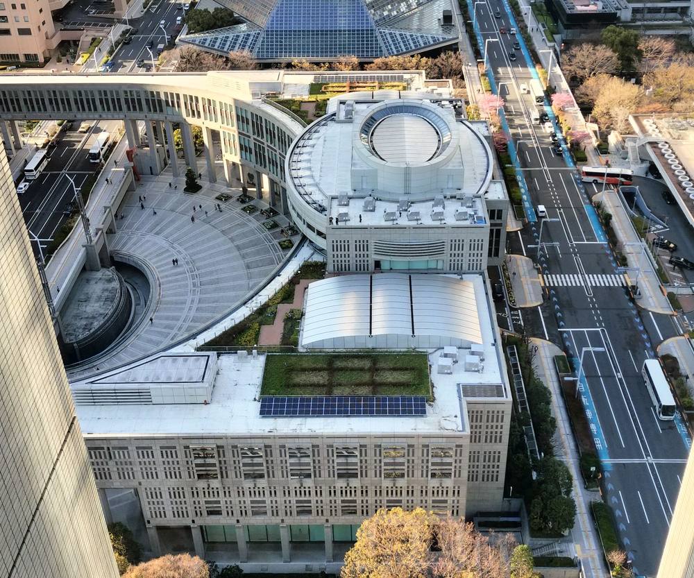 白天從高樓往下俯瞰，中央是屋頂有圓形天窗的都議會議事堂，左側是鋪著同心圓石磚的半圓形廣場，廣場被議事堂的弧形樓層環抱，左緣切進一棟高樓的牆面，右側是多線道馬路
