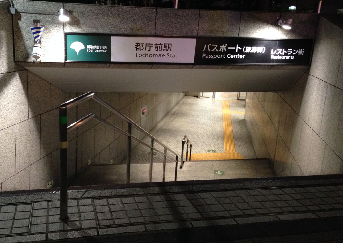 夜間往下通往地下的石材樓梯口，上方橫掛的招牌左邊是都營地下鐵的綠色標誌與「都庁前駅 Tochomae Sta.」，右邊是黑底的「パスポート（旅券課）Passport Center」「レストラン街 Restaurants」，樓梯中間有不鏽鋼扶手，底下是有黃色導盲磚的通道