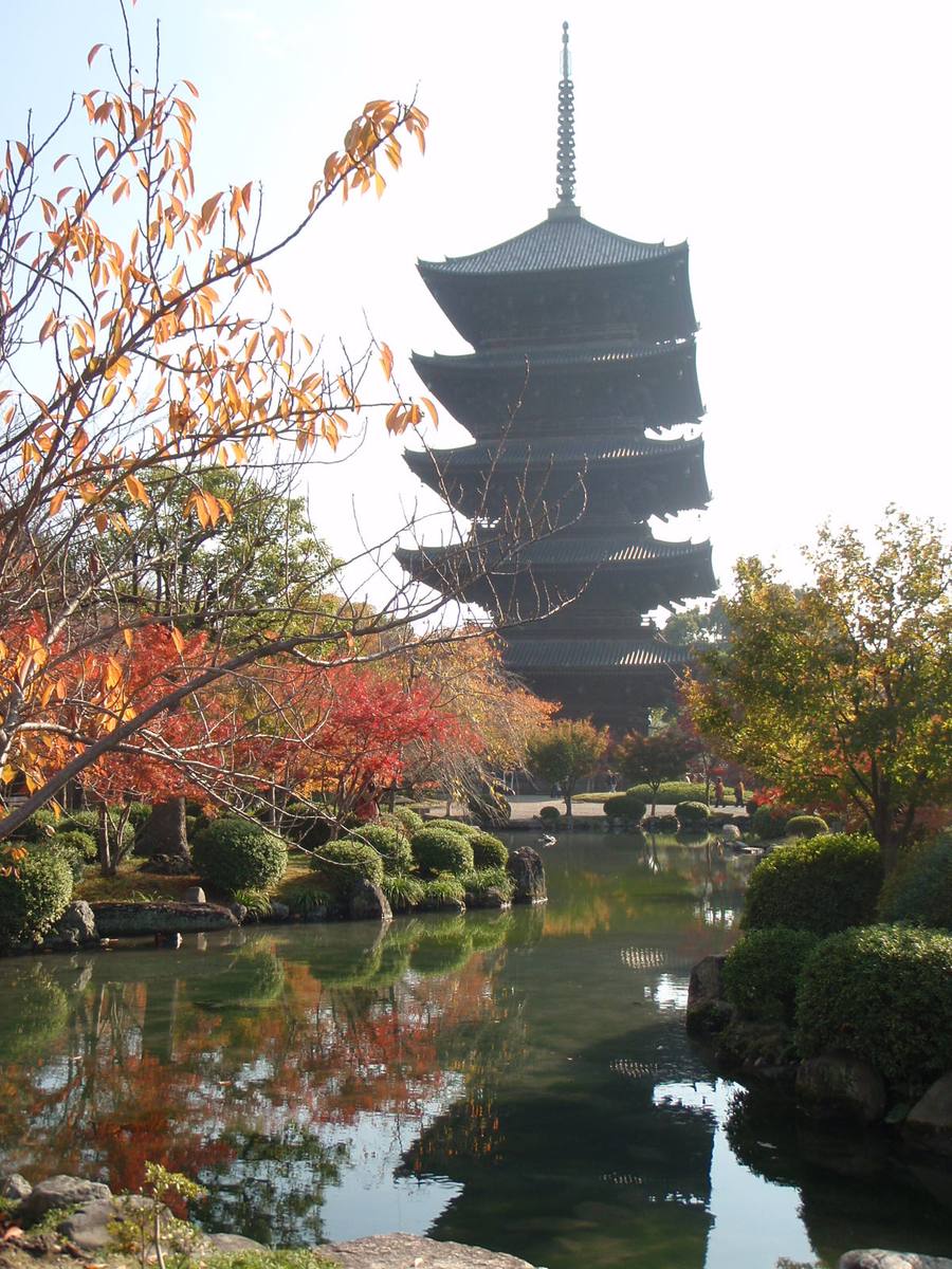 白天的東寺五重塔與瓢箪池:深色木造塔身立在池後,池邊是紅葉與修成圓形的灌木,前景有橘黃色的枝葉,水面映著樹影