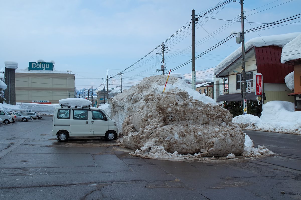 十日町市區一處路口,馬路中央堆著一座髒雪堆,高度明顯超過旁邊那台白色輕型廂型車的車頂兩倍,雪堆上插著一根紅白相間的標桿。左後方是掛著「Daiyu」招牌的賣場,右側日式店家的屋頂與雨庇上壓著厚厚的雪,店門旁的雪也堆到窗台高度。柏油路面是被除雪後的濕黑色