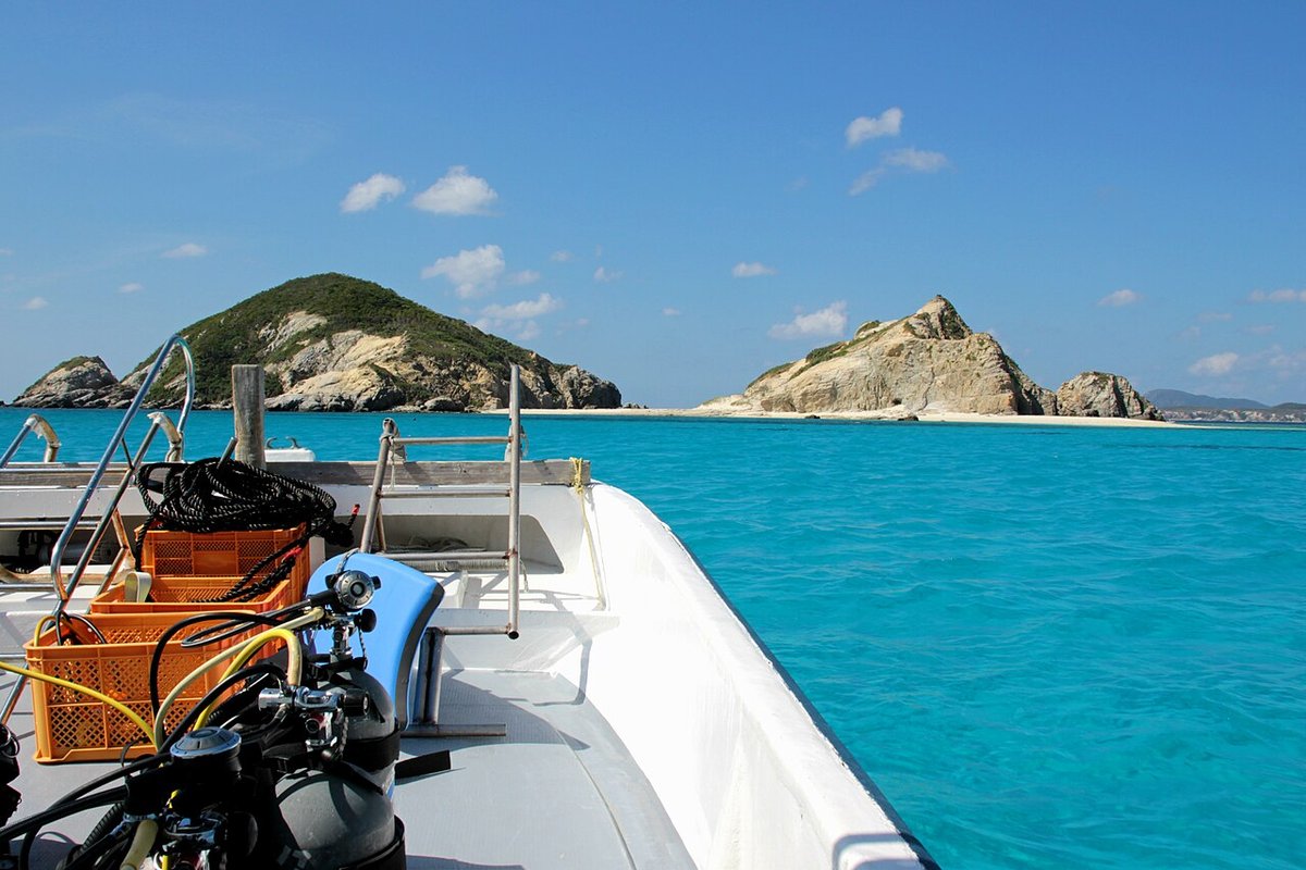 View forward from the bow of a small boat, scuba cylinders, regulators and orange crates on the deck, the bow pointing at two rocky islets with a white beach and glowing pale green water between them